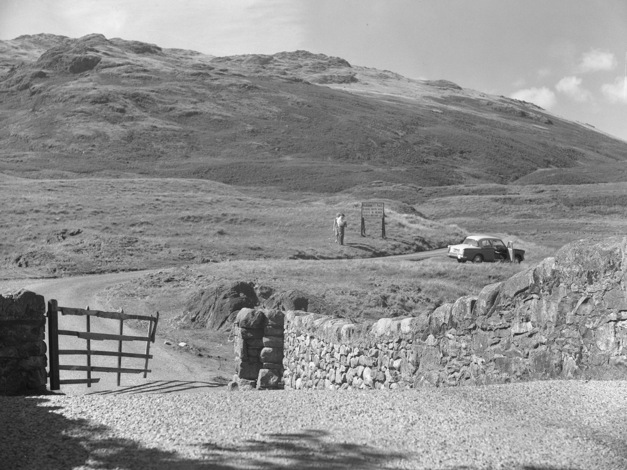 A Hard Knott hágón áthaladó út, két ember áll egy útjelző tábla mellett és egy parkoló autó a füves szélén. alkotó: Joseph Hardman