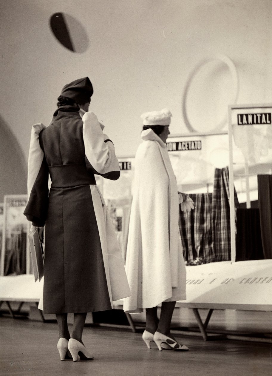 A hátulról látható két modell a "Lamma" couture ház alkotásait viseli. Az egyik modell két tónusú felöltőt visel, derékban öblítéssel. A másik modell teljes köpenyt visel. Az azonos színű kalapok minden együttest kiegészítenek. (f/w fotó) alkotó: Luis Bogino