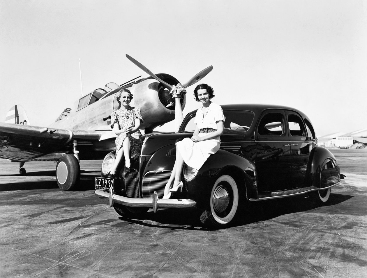 Kalifornia: 1939 Két nő ül egy 1939-es Lincoln Zephyr szedán sárvédőin egy repülőtéren, mögöttük egy hajtóműves gép parkol (f/w fotó) alkotó: Unbekannt