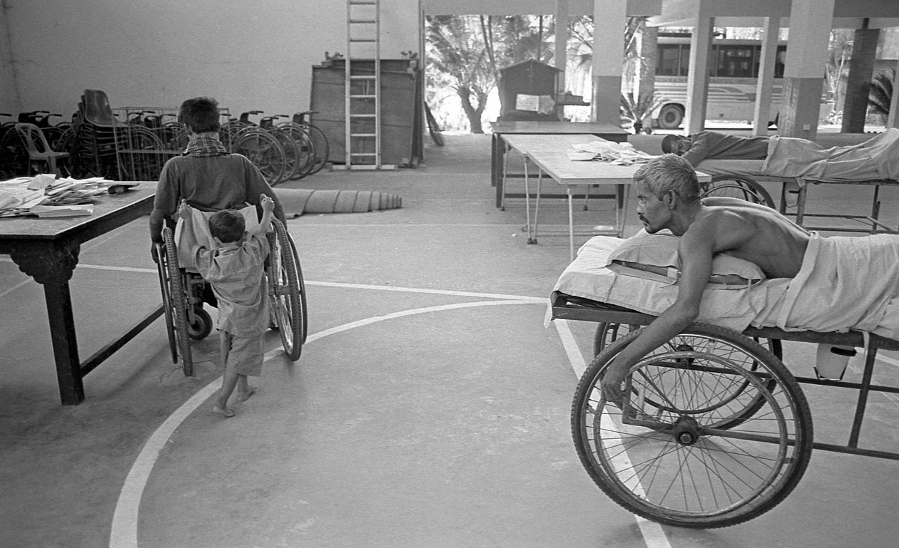 Egy mozgássérült férfit tolószékben a fia visz; Egy másik ember, aki ugyanilyen helyzetben van, nézi a Bénultak Rehabilitációs Központjában, a CRP Savarban (Dhaka, Banglades). 2005. alkotó: Unbekannt Unbekannt