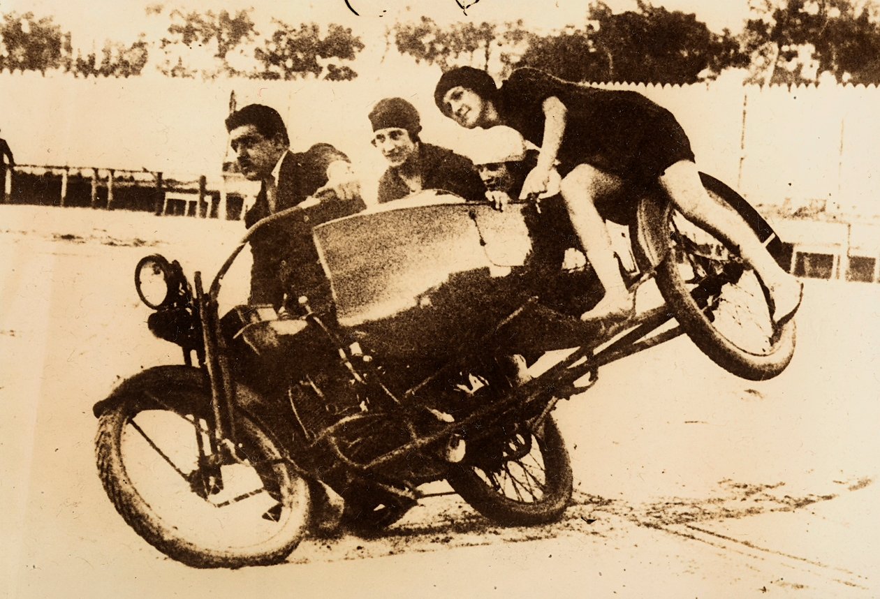 Egy férfi egy motorkerékpár vezetésével mutatkozik be, amelynek oldalkocsija három fiatal nőt szállít. Az oldalkocsi felemelkedik a földről, amikor kanyarodnak. alkotó: Unknown photographer