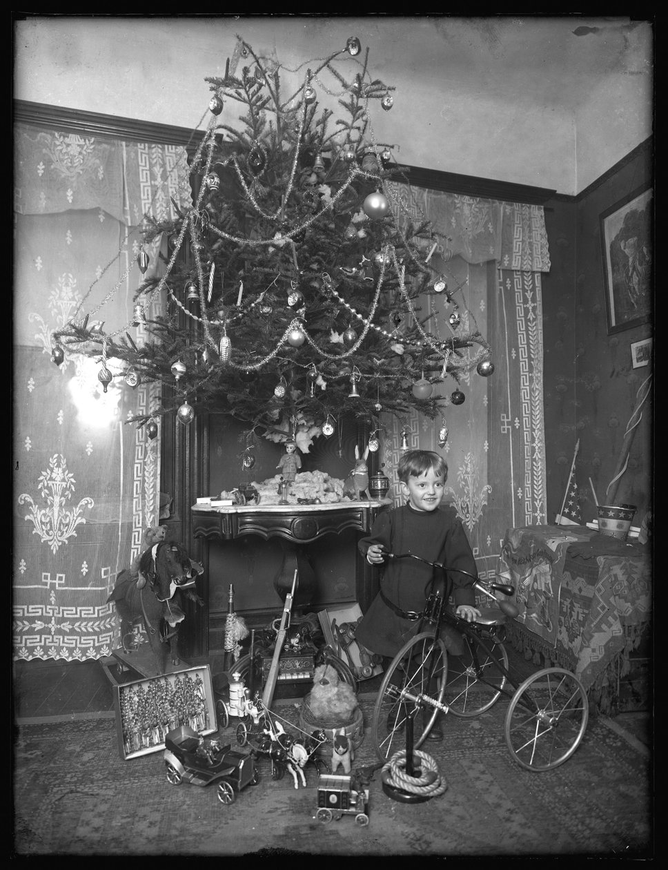 Seymour fiú triciklivel pózol a karácsonyfa mellett a szalonban, 1912 karácsonya alkotó: William Davis Hassler