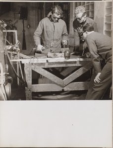 [Három szemüveges férfi hegeszt egy fémdarabot egy asztalon]
