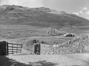 A Hard Knott hágón áthaladó út, két ember áll egy útjelző tábla mellett és egy parkoló autó a füves szélén. alkotó: Joseph Hardman