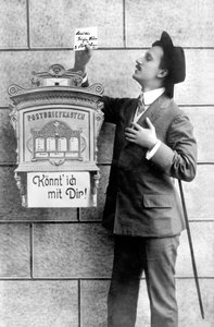 Egy elegáns férfi öltönyben, kalapban és botban posztol egy üzenetet a postaládába. Németország 1920 körül Történelmi fénykép, férfi egy levélszekrény mellett, 1920 körül alkotó: Unbekannt