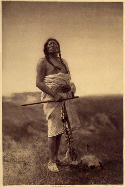 Férfi orvos. Valószínűleg sziúk, tintahal a kezében, és bivalykoponya a lába. A fénykép Edward S. Curtis észak-amerikai indiánokról szóló enciklopédiájának (1868-1952) 3. kötetéből készült, Theodore Roosevelt előszavával. 1907 és 1930 között 20 kötet jelen alkotó: Edward Sheriff Curtis