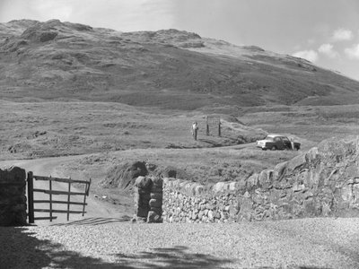 A Hard Knott hágón áthaladó út, két ember áll egy útjelző tábla mellett és egy parkoló autó a füves szélén. alkotó: Joseph Hardman