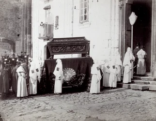 A nápolyi San Francesco Kongregáció temetési menete. Az előtérben, egy templom előtt számos fehér tunikás és csuklyás „társi testvér” kíséri a hímzett ruhával díszített katafalkot, amelyre a koporsót helyezik.