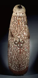 Ovális Asmat pajzs, domborműben faragott görgetős díszítéssel, erősen stilizált figurákkal, fonott bojtokkal a szegélyig (fa, raffia és toll)