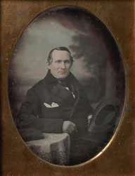 Kabátot és kesztyűt viselő férfi, kalapot tart, festett kültéri háttér előtt ül, 1840-es évek.