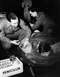 Második világháború (1939-1945) – II. világháború (II vagy WW2): Luxemburg 1945. február: Az Egyesült Államok hadseregének evakuációs kórházában egy orvos (ápolónő) penicillint ad be egy sérült katonának, hogy elkerülje a fertőzést, miközben egy nővér kész