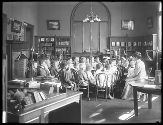 A gyerekek egy nőt hallgatnak a Queens Borough Public Library egy azonosítatlan fiókjának olvasótermében, 1910 körül