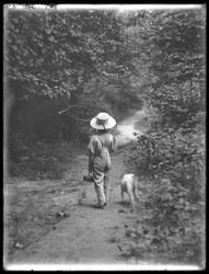 William Gray Hassler szalmakalapban és kezeslábasban horgászbottal és kosárral, sétál egy ösvényen az erdőben egy kis kutyával (Bessie), 1911. augusztus 20.